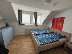 A bedroom with a sloped ceiling, a bed with a patterned bedspread, a dresser, a radiator, and a window with curtains.