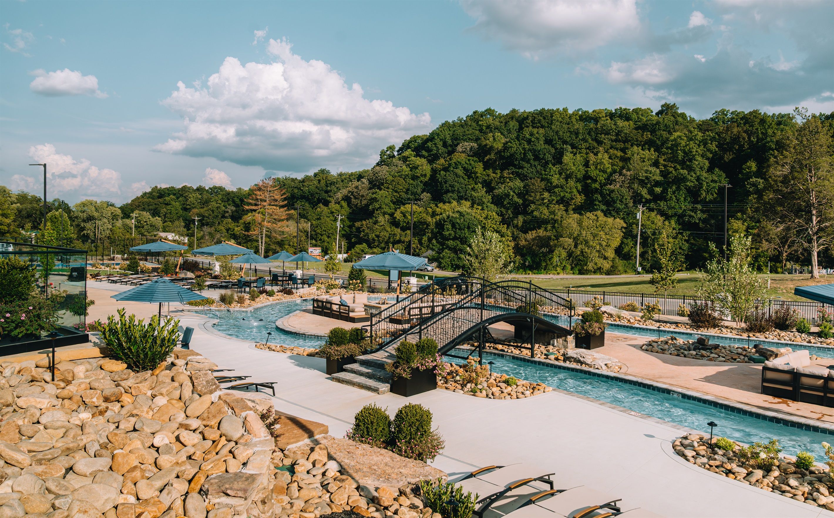 A pool area with a bridge, lounge chairs, and a view of a forested hill.