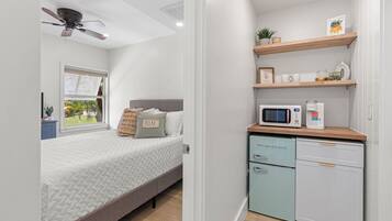 A compact bedroom with a bed, a ceiling fan, a window with blinds, a kitchenette with a microwave, and a small cabinet.