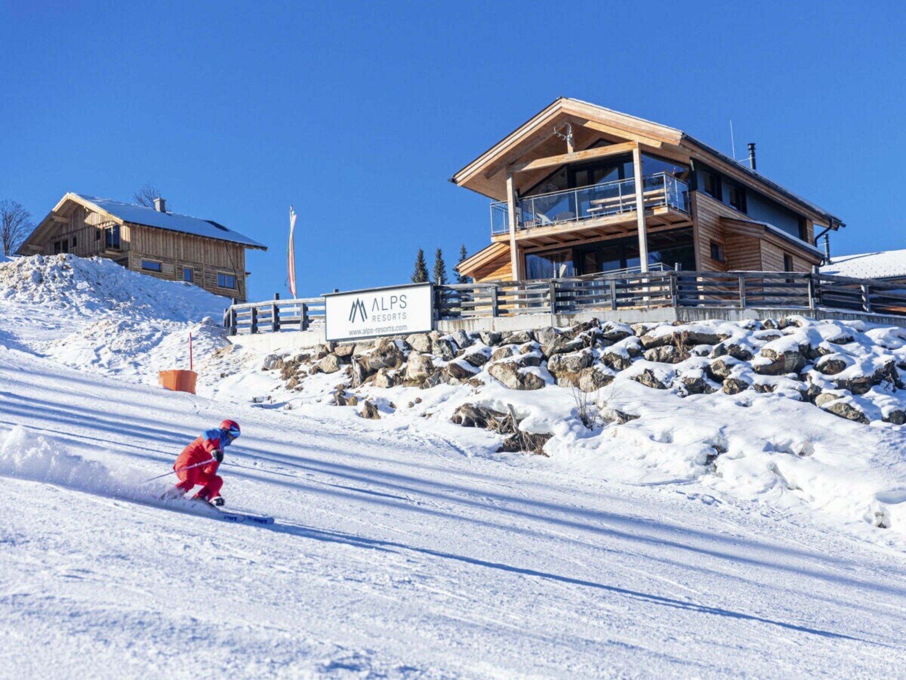 Ein Skigebiet mit einer Holzhütte, einem Schild für ALPS RESORTS und einem Skifahrer in einem verschneiten Hang.