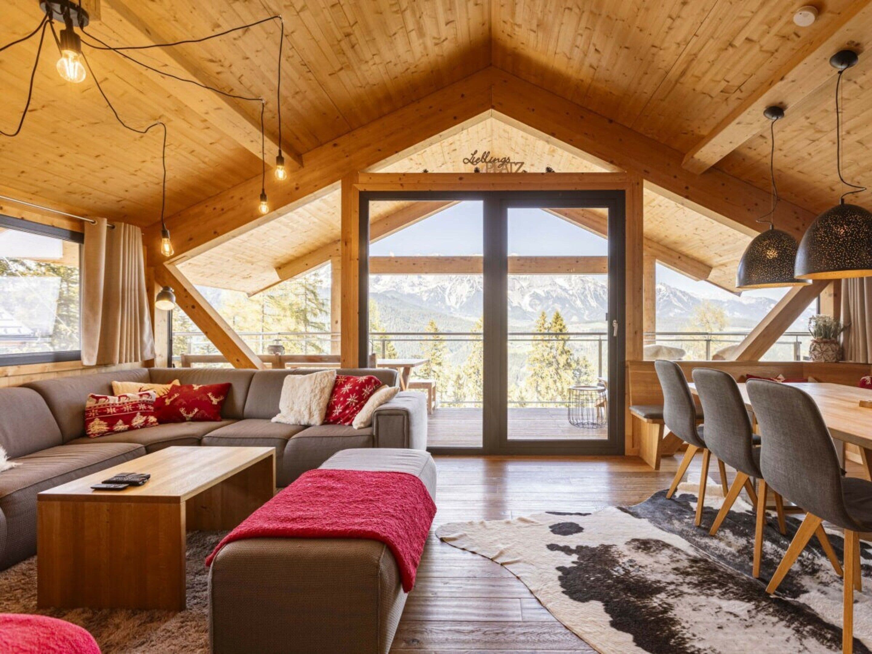 Una habitación acogedora estilo loft con techo de madera, un sofá, una mesa de comedor con sillas y vistas a montañas a través de un amplio ventanal.