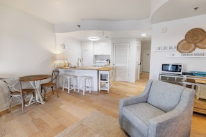 A compact living space with a kitchenette, a dining area with a wooden table and chairs, a grey armchair, and a TV. - The View at Carillon - Lake & Gulf Views + Resort Amenities (Panama City Beach)