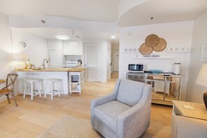 A modern kitchen with white cabinetry, a dining area with a wooden table and white chairs, and a living room with a grey armchair and a coffee machine. - The View at Carillon - Lake & Gulf Views + Resort Amenities (Panama City Beach)