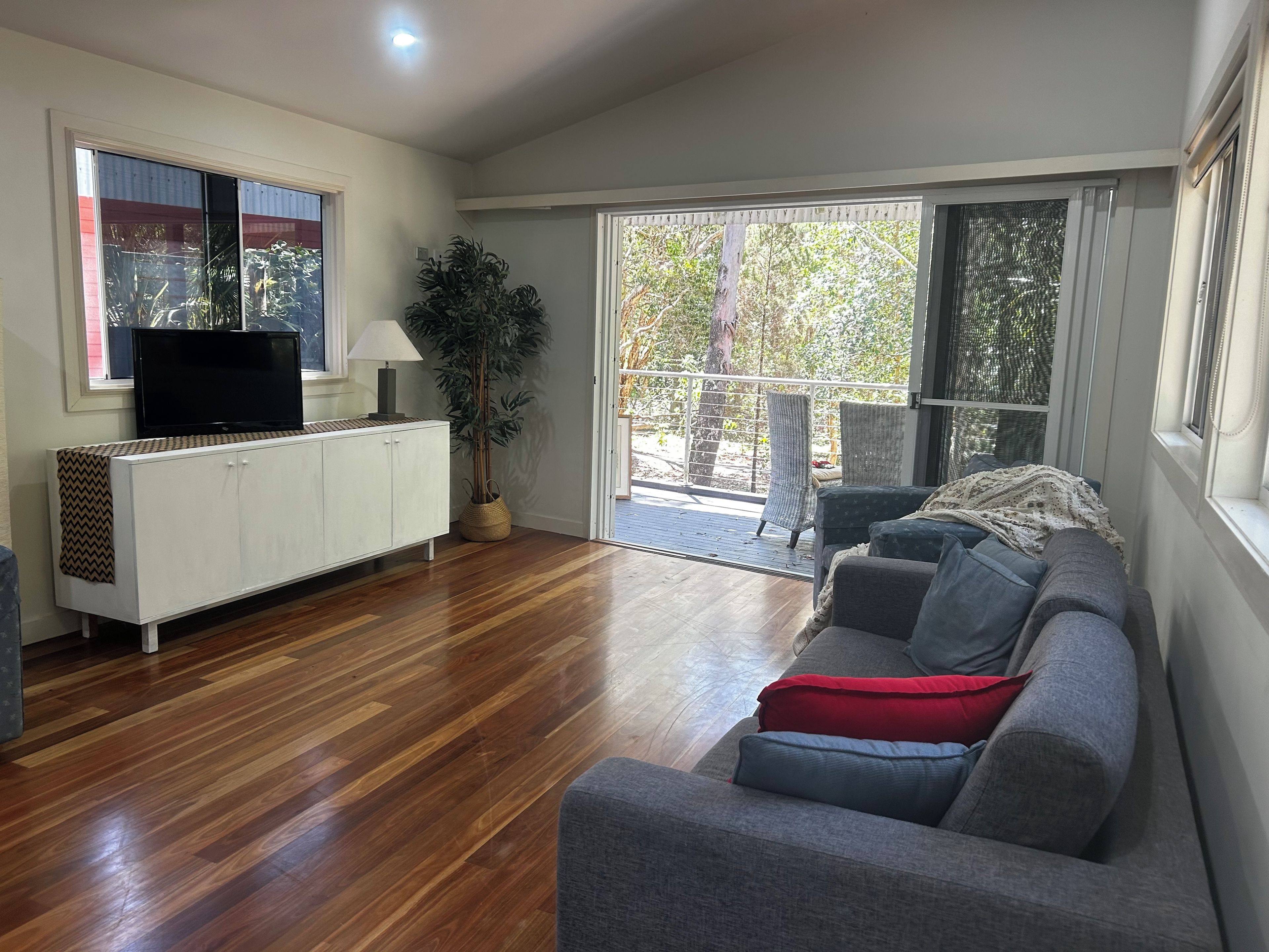Ein Wohnzimmer mit grauem Sofa, einem Fernseher auf einem weißen Schrank, einem Blumentopf und einer Glastür, die zu einem Außenbereich führt.