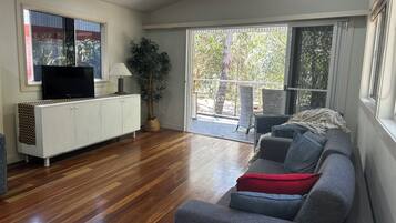 A living room with a grey sofa, a TV on a white cabinet, a potted plant, and a sliding glass door leading to an outdoor area.