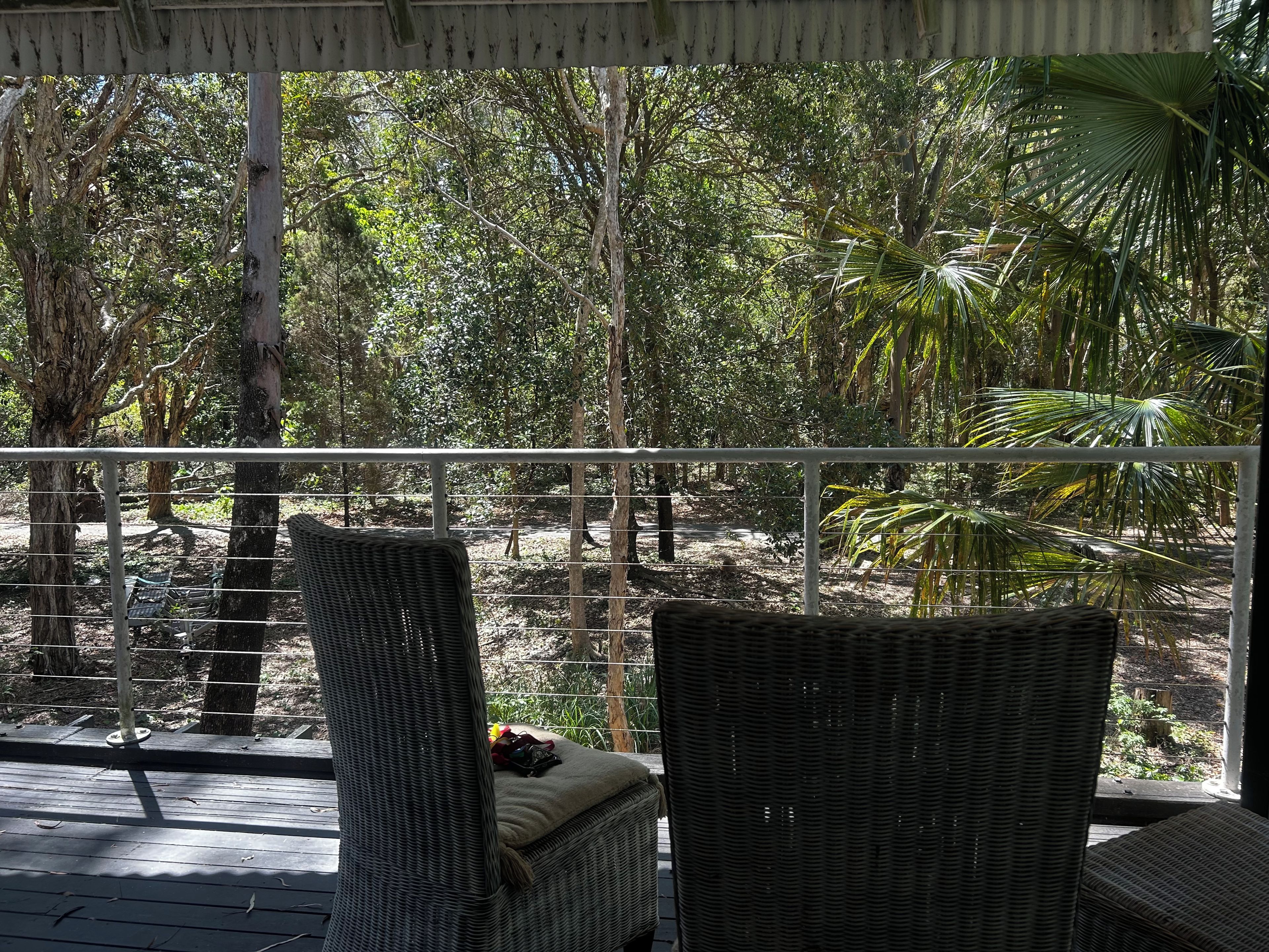 A covered outdoor seating area with wicker chairs and a bench, overlooking a natural landscape with trees and a pathway.