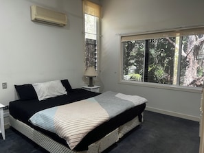 A bedroom with a bed, a nightstand, a lamp, a window with a view of trees, and an air conditioning unit.