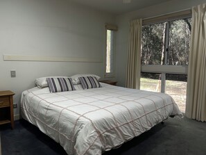 A bedroom with a large bed, a nightstand, a window with curtains, and a view of trees outside.