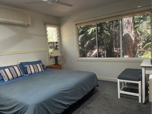 Ein Schlafzimmer mit einem Bett, einem Nachttisch, einer Lampe, einem Fenster mit Blick auf Bäume und einem Deckenventilator.