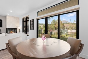 Ein modernes Wohnzimmer mit Kamin, einem runden Esstisch mit Stühlen und großen Fenstern mit Blick auf eine gebirgige Landschaft.
