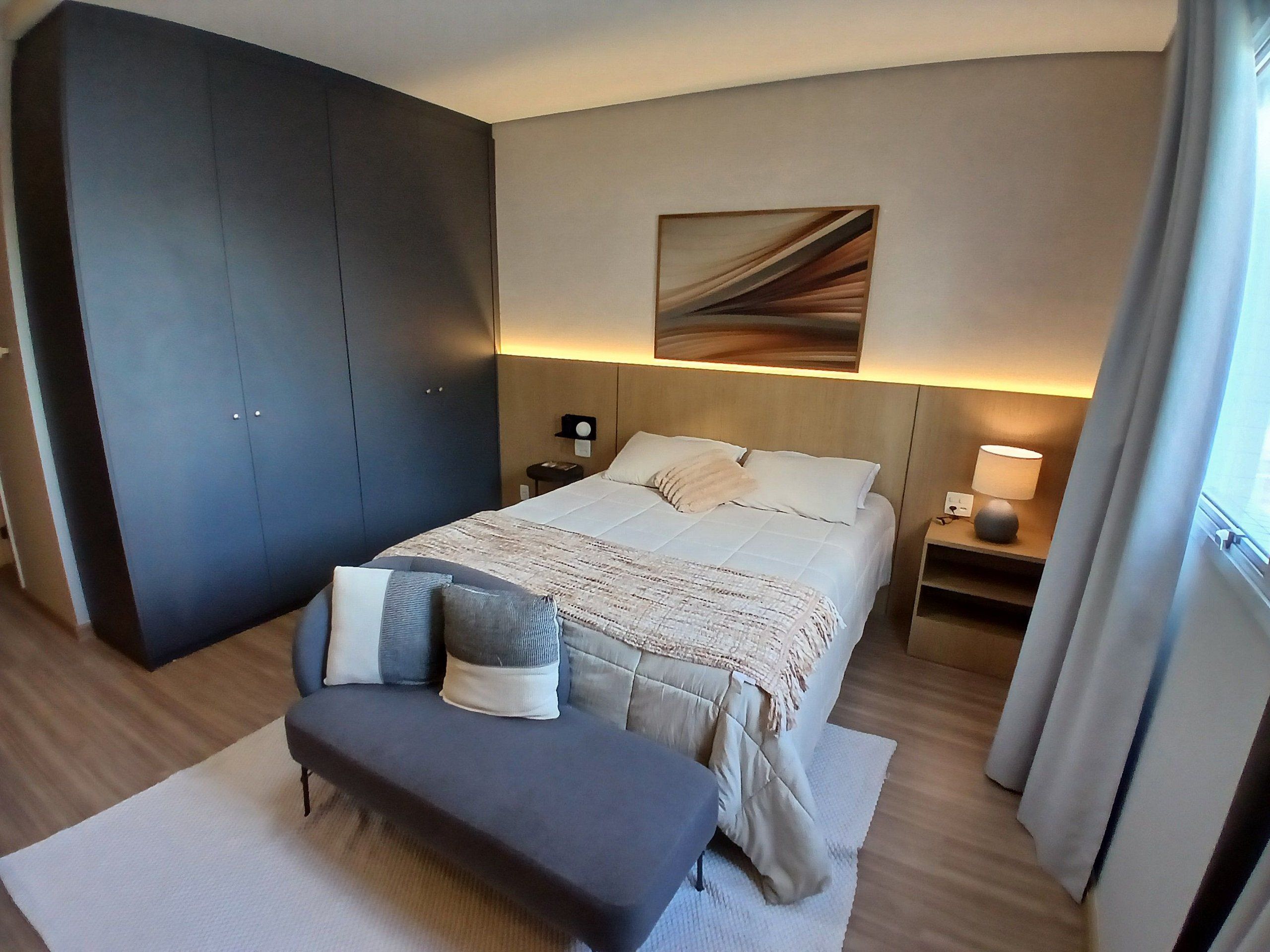 Una habitación de hotel con cama, mesitas de noche, un banco, un armario grande y un cuadro en la pared.