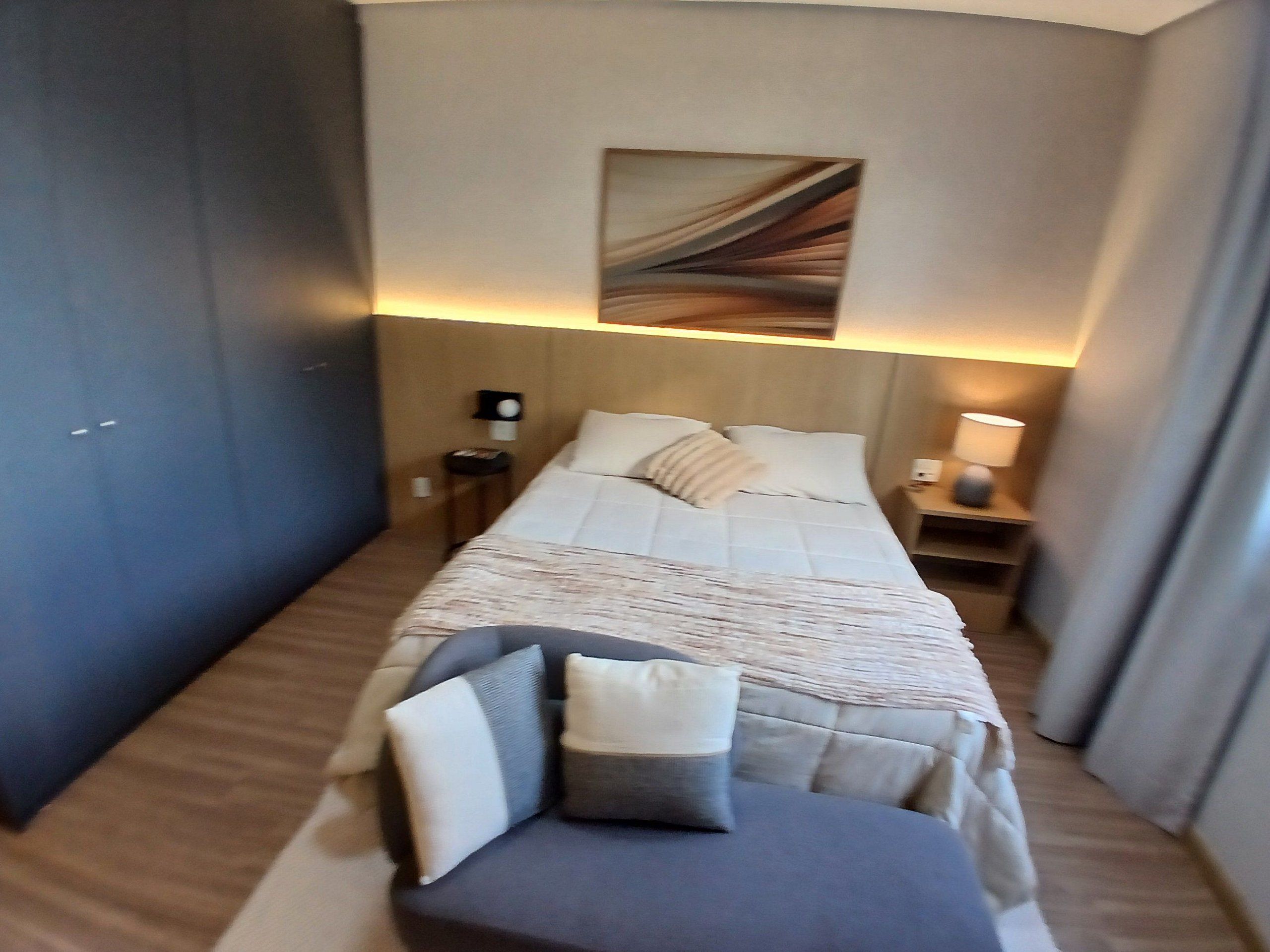 Habitación de hotel con una cama, una mesita de noche con lámpara, un pequeño atril y una obra de arte en la pared.