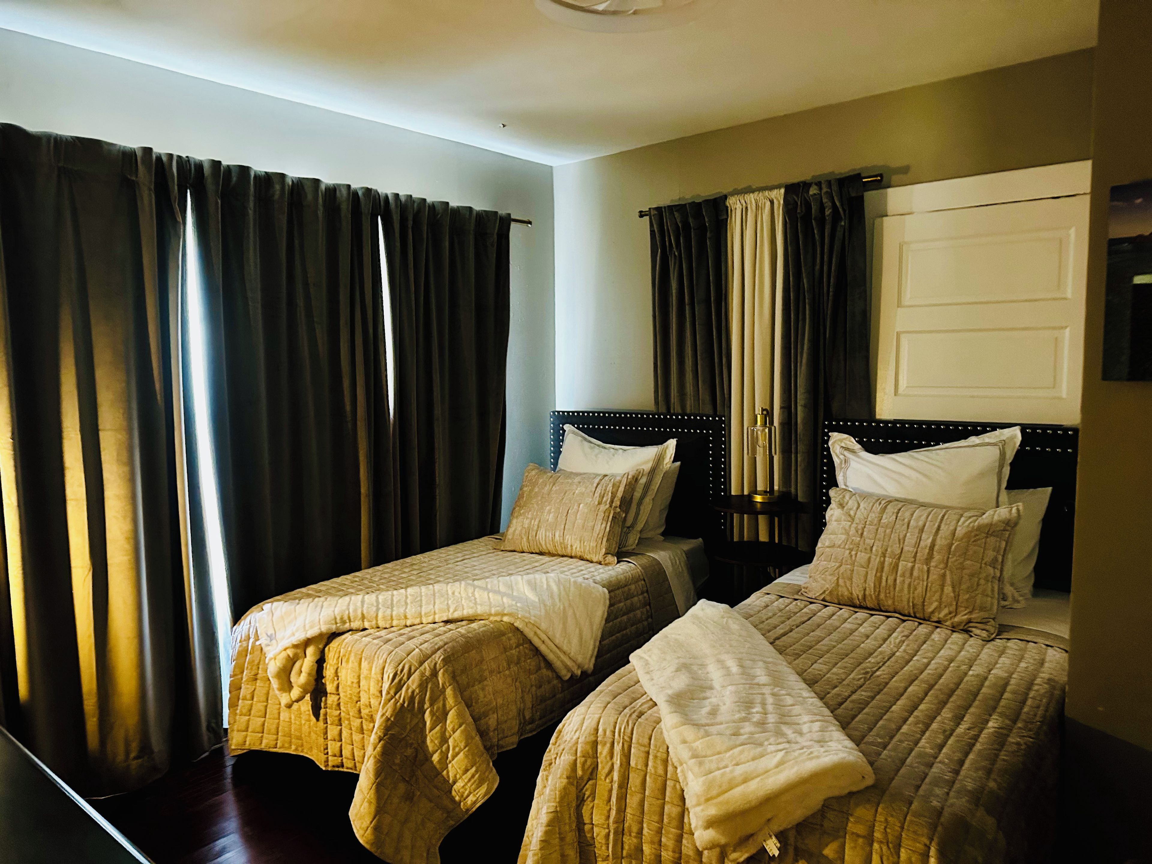 A hotel room with two beds, dark curtains, a bedside table with a lamp, and a door.