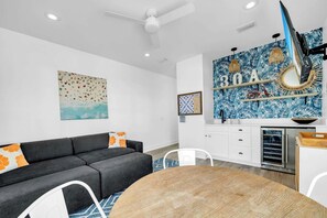 A modern living room with a gray sofa, a round wooden table, and a kitchen area with a blue tropical wallpaper. - Good Day Sunshine by Stay on 30a (Seacrest Beach)