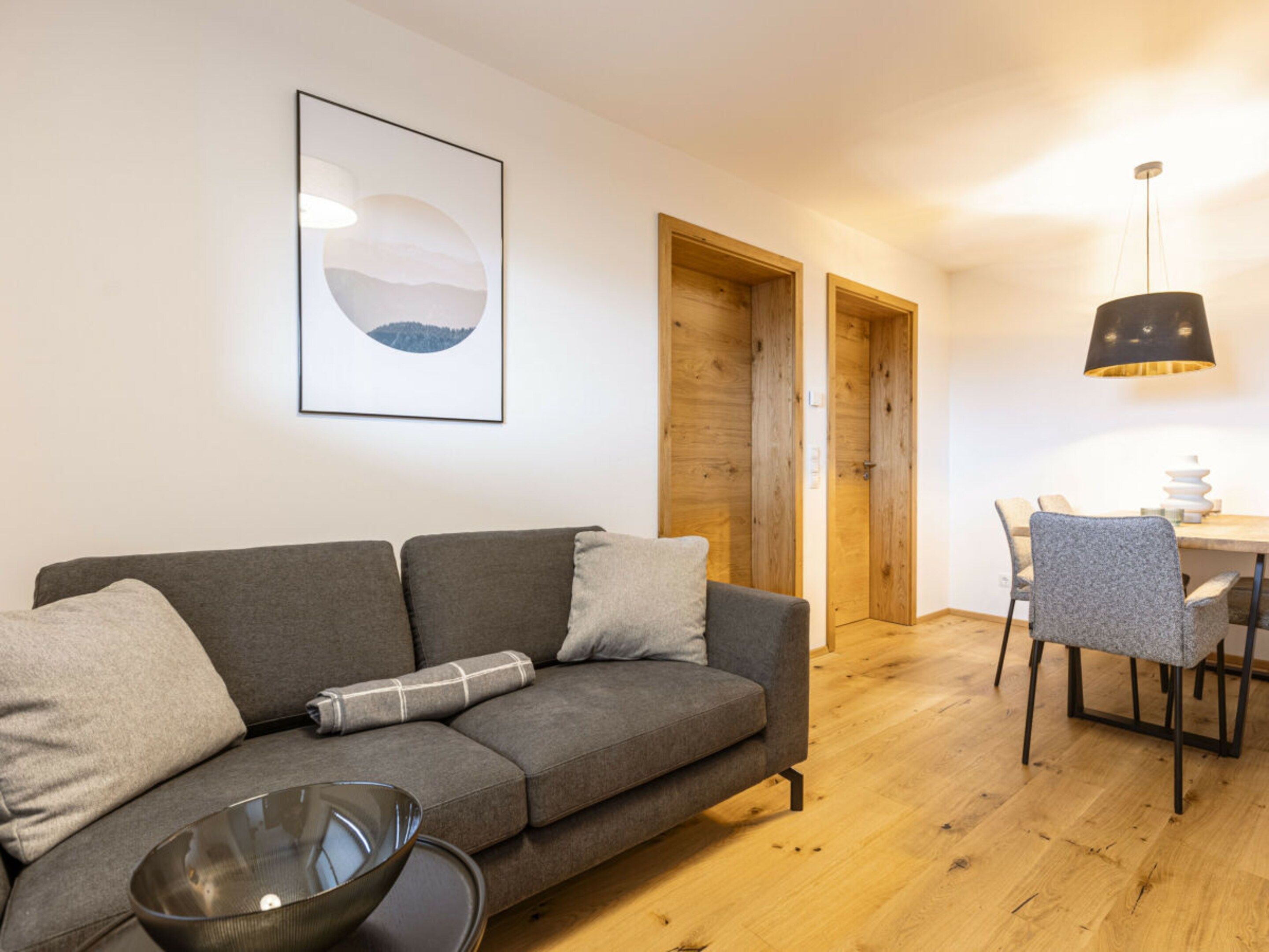 Ein modernes Wohnzimmer mit einem grauen Sofa, einem Esstisch mit Stühlen, Holztüren und einer Pendelleuchte.