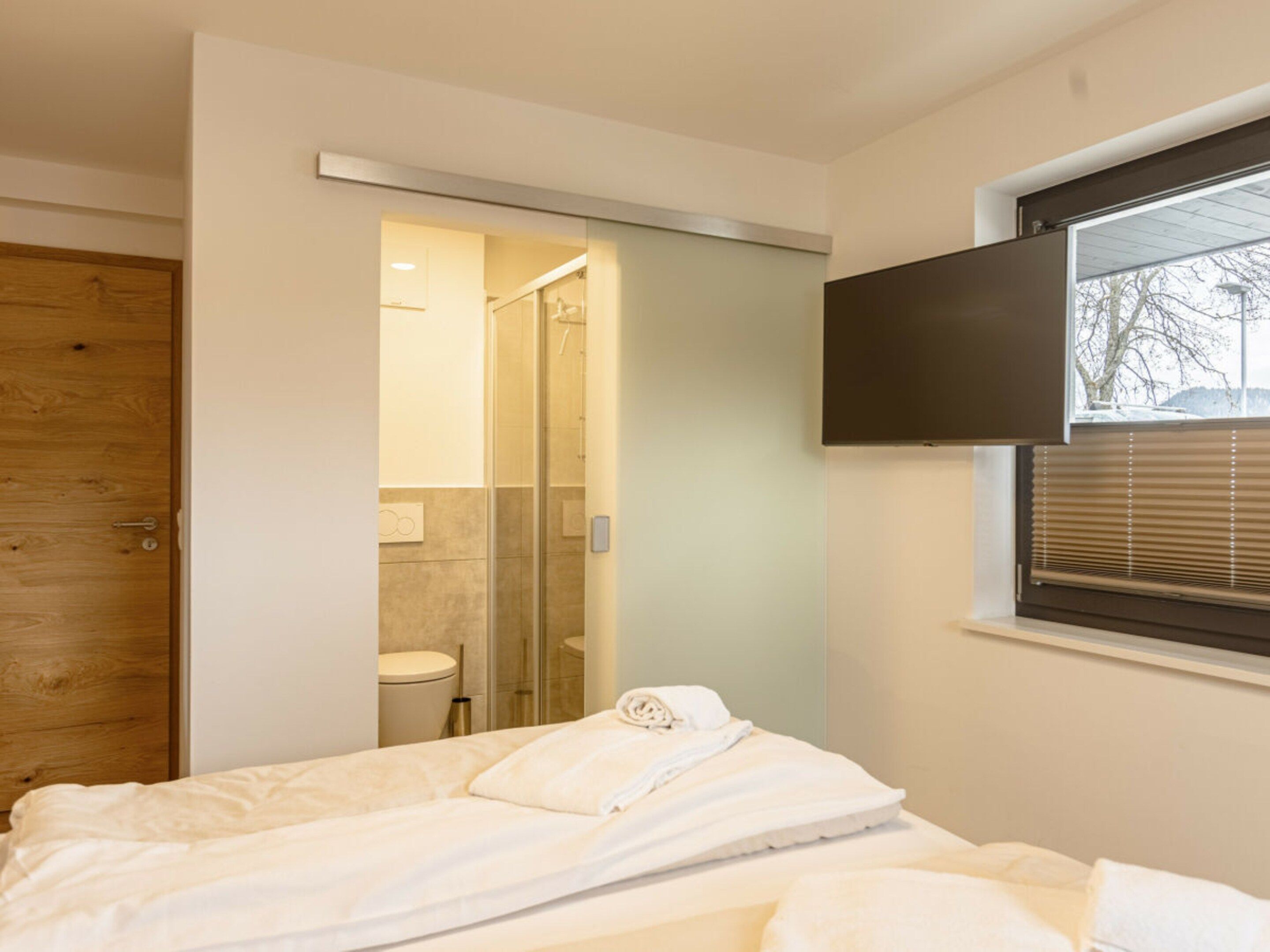 Un dormitorio con cama, puerta de madera, baño con ducha y ventana con persianas.