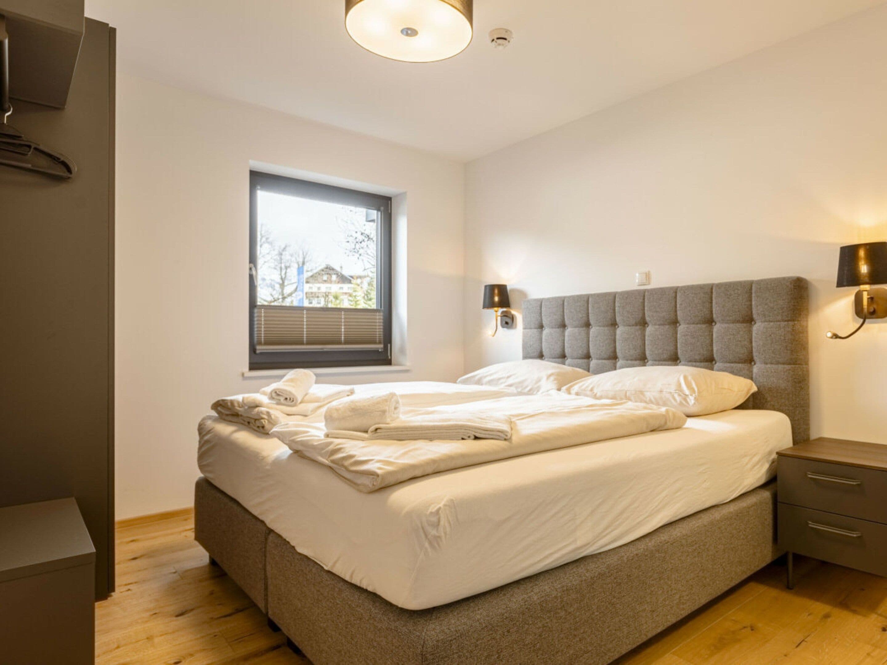 A modern bedroom with a large bed, bedside tables, a window with blinds, and a view of a building outside.