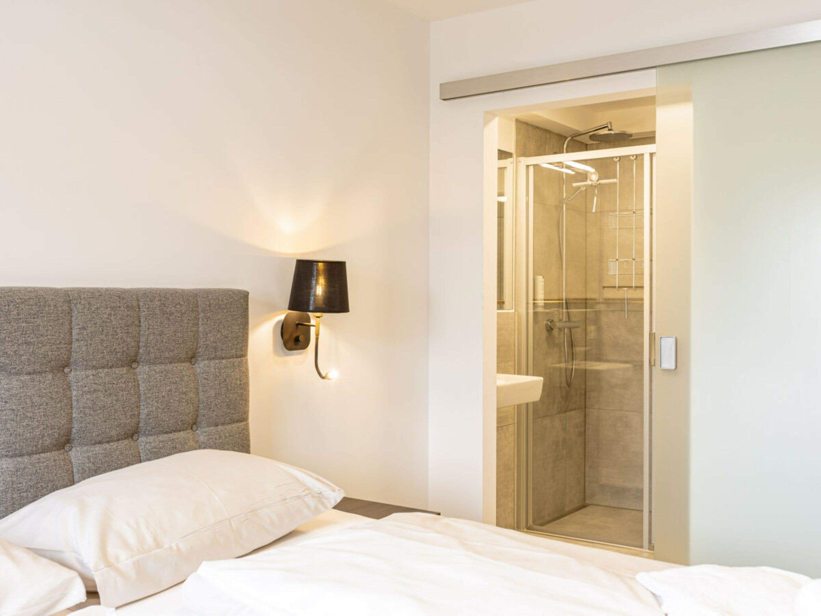 A bedroom with a bed, a wall-mounted lamp, and a glass-enclosed shower area.