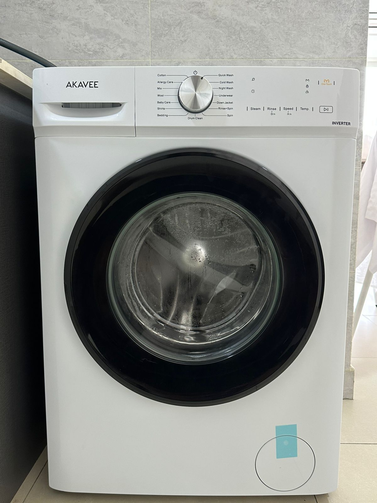 A front view of a white Akavee washing machine with a control panel, a circular door, and a small blue indicator light.
