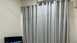 Ein Wohnzimmer mit grauem Sofa, einem Fernseher auf einem Schrank und einem Fenster mit grauen Vorhängen.
