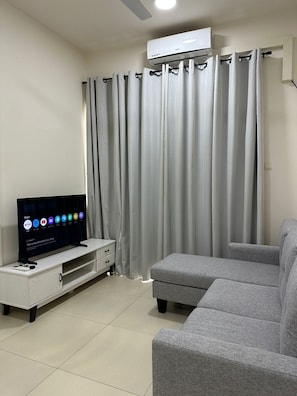 Una sala de estar con un sofá gris, un televisor sobre un mueble bajo y una ventana con cortinas grises.