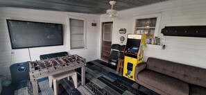 A room with a foosball table, a sofa, a wall-mounted TV, and a Pac-Man arcade machine.