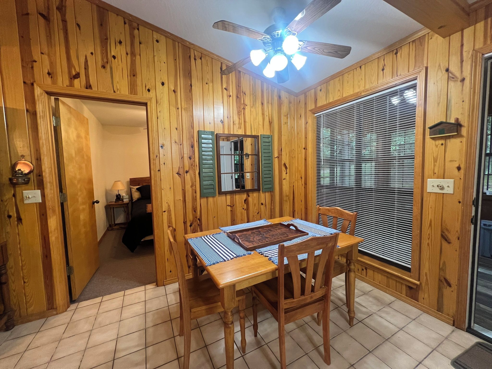 A wooden-paneled room with a dining table, chairs, a ceiling fan, and a window with blinds.