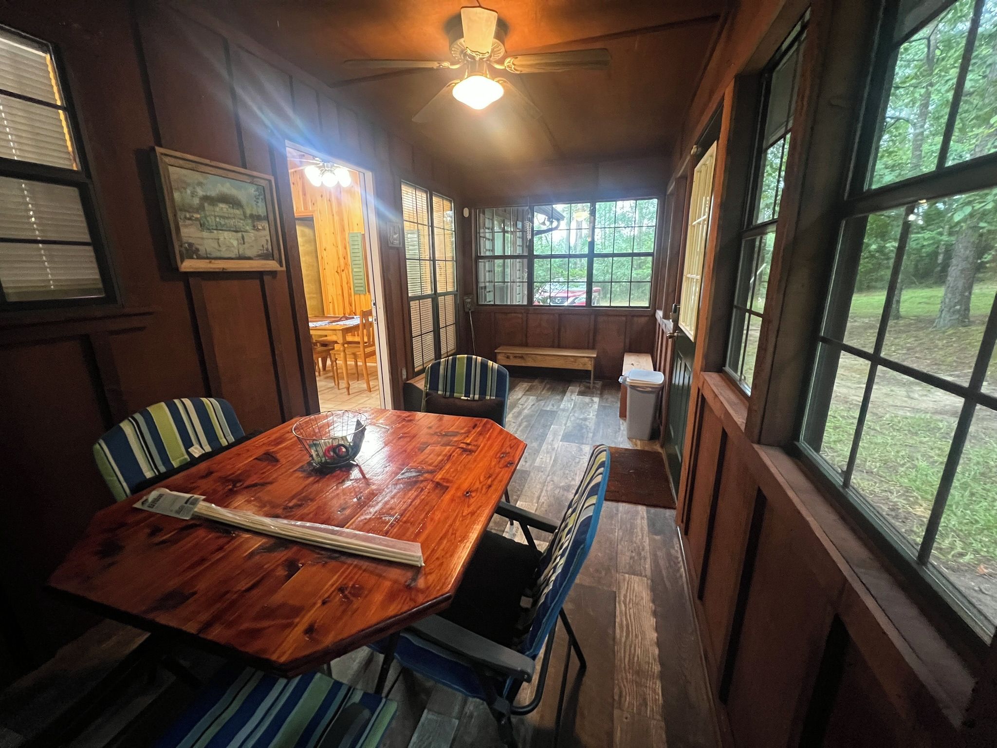 A wooden dining table with chairs, a ceiling fan, and large windows with a view of the outdoors.