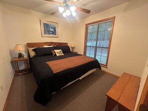 A bedroom with a bed, a ceiling fan, a window with blinds, a bedside table with a lamp, and a framed picture on the wall.