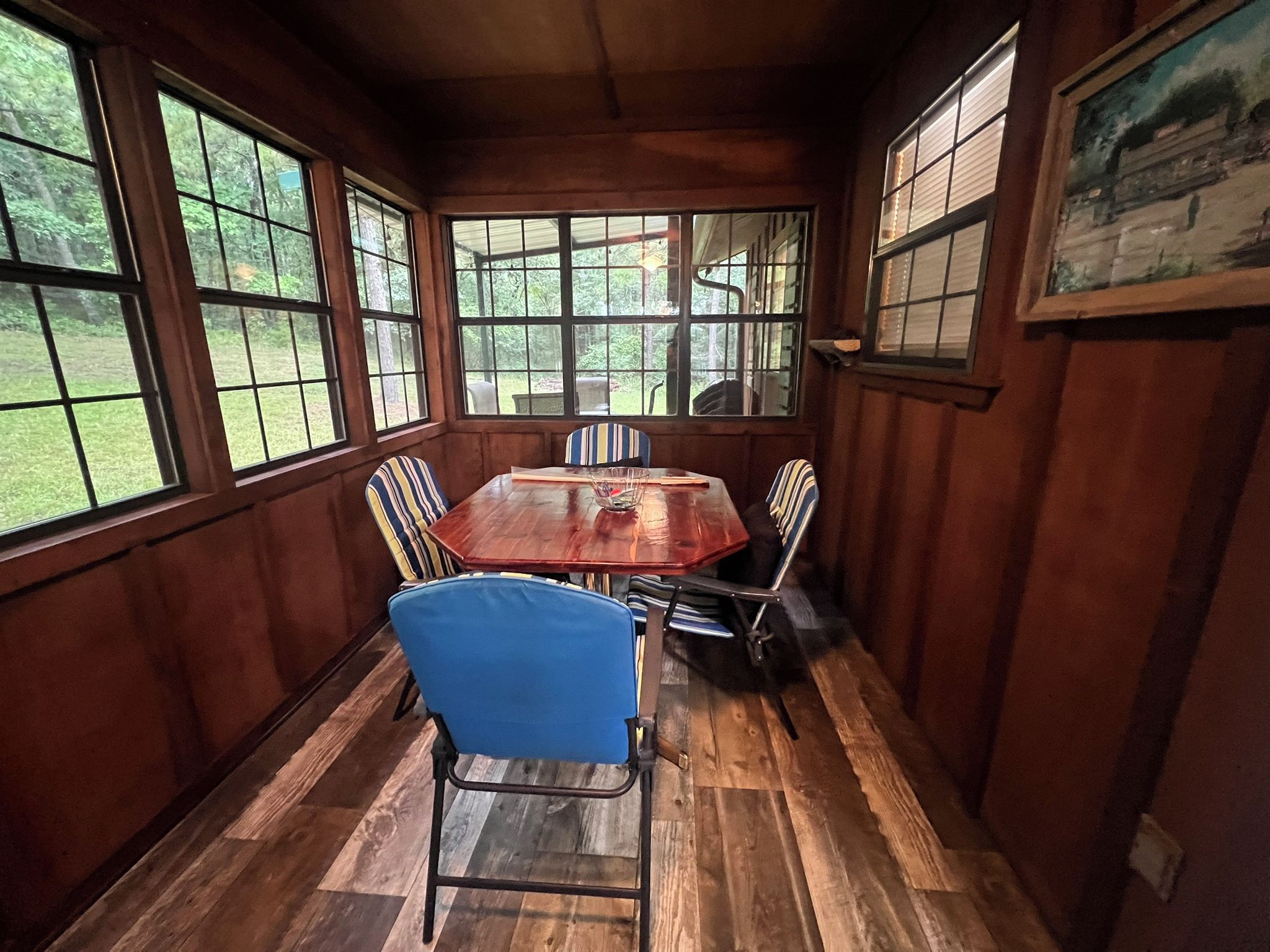 A dining room with a wooden table and chairs, large windows, and a painting on the wall.