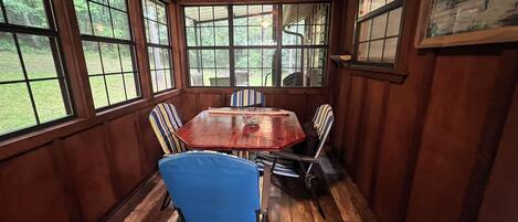 A dining room with a wooden table and chairs, large windows, and a painting on the wall.