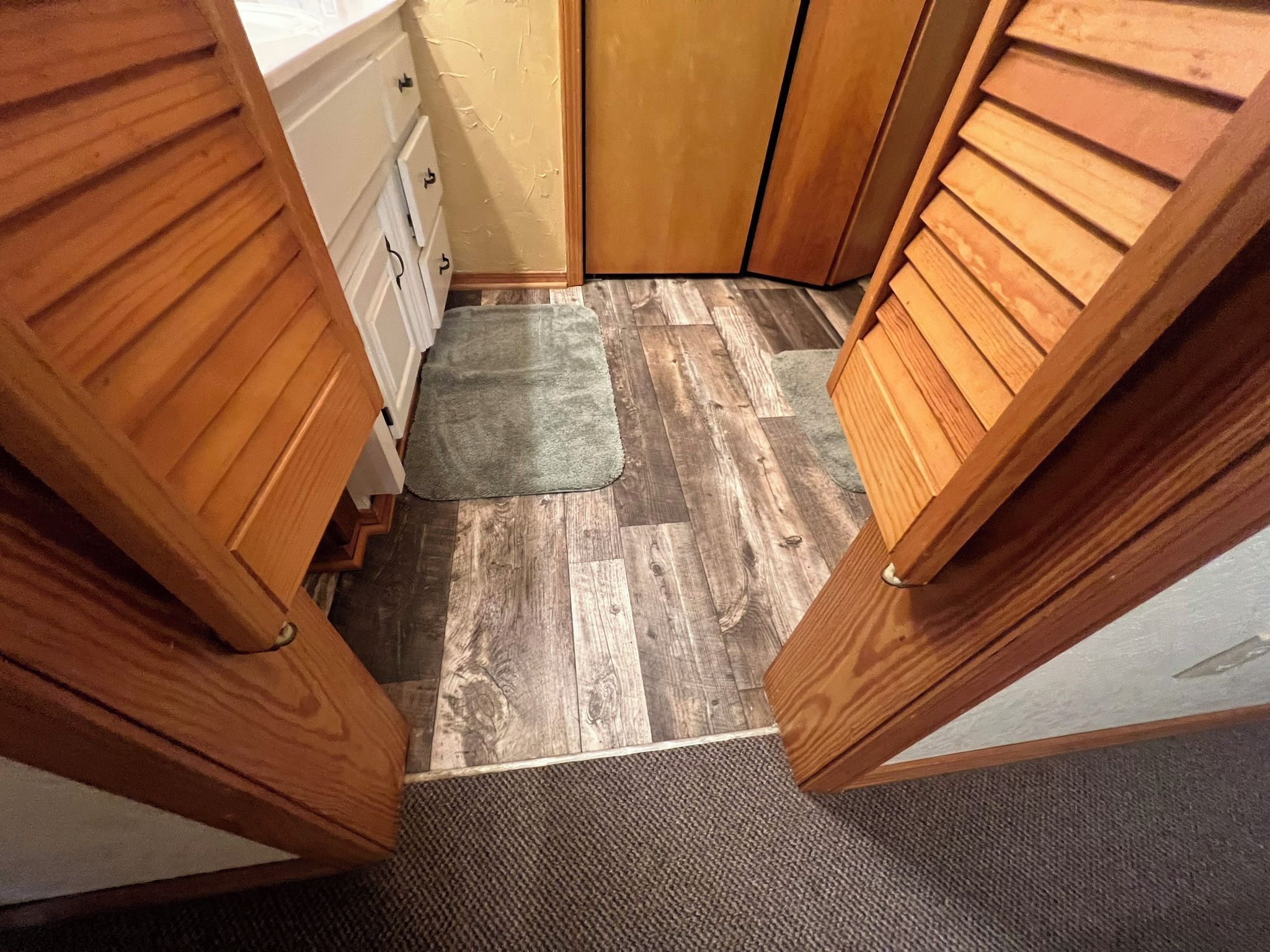 A narrow hallway with wooden doors, a gray mat, and a wooden floor.