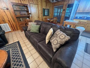 A living room with a dark brown leather sofa, patterned cushions, a tiled floor, wooden walls, and a dining area with a table and chairs.