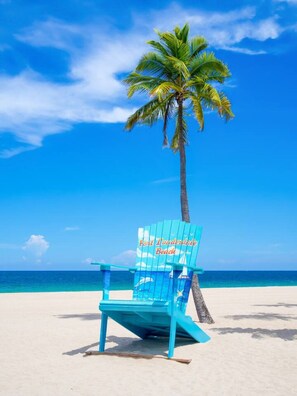 A turquoise beach chair with 'Fort Lauderdale Beach' written on it, positioned on a sandy beach with a palm tree and clear blue sky in the background. - W Penthouse | Ocean Views | Rooftop Pool (Fort Lauderdale)