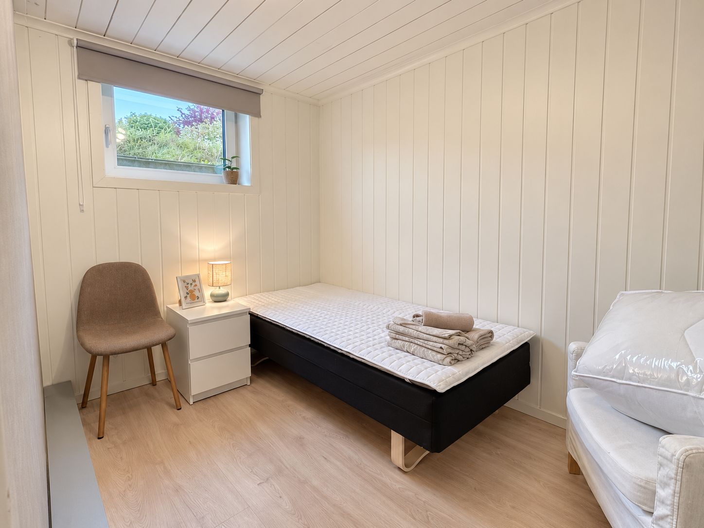 A small, neatly arranged bedroom with a single bed, a chair, a bedside table with a lamp, and a window with a view of greenery.