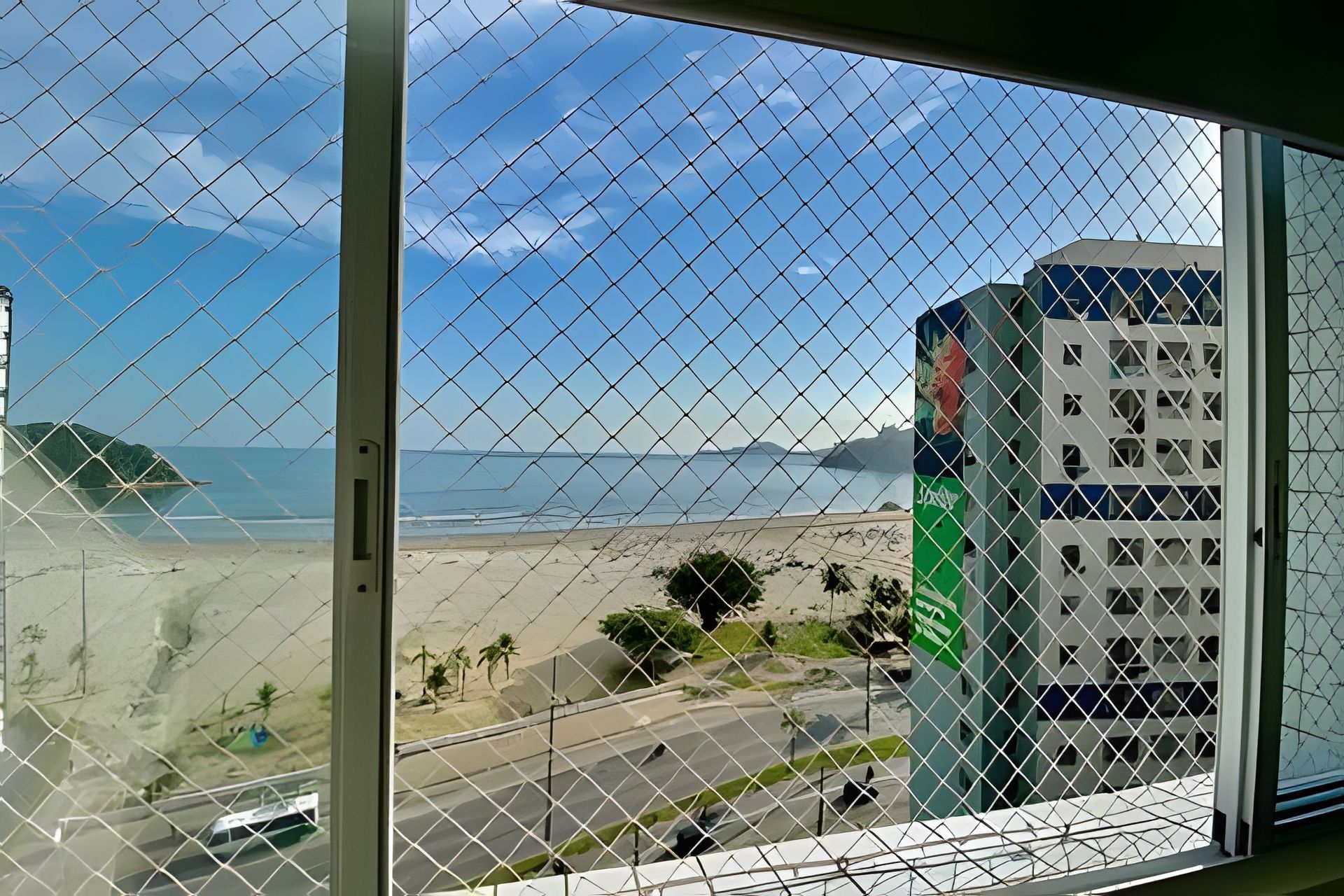 Vista a través de una cerca de alambre de una playa, una carretera y un edificio con fachada verde y blanca.
