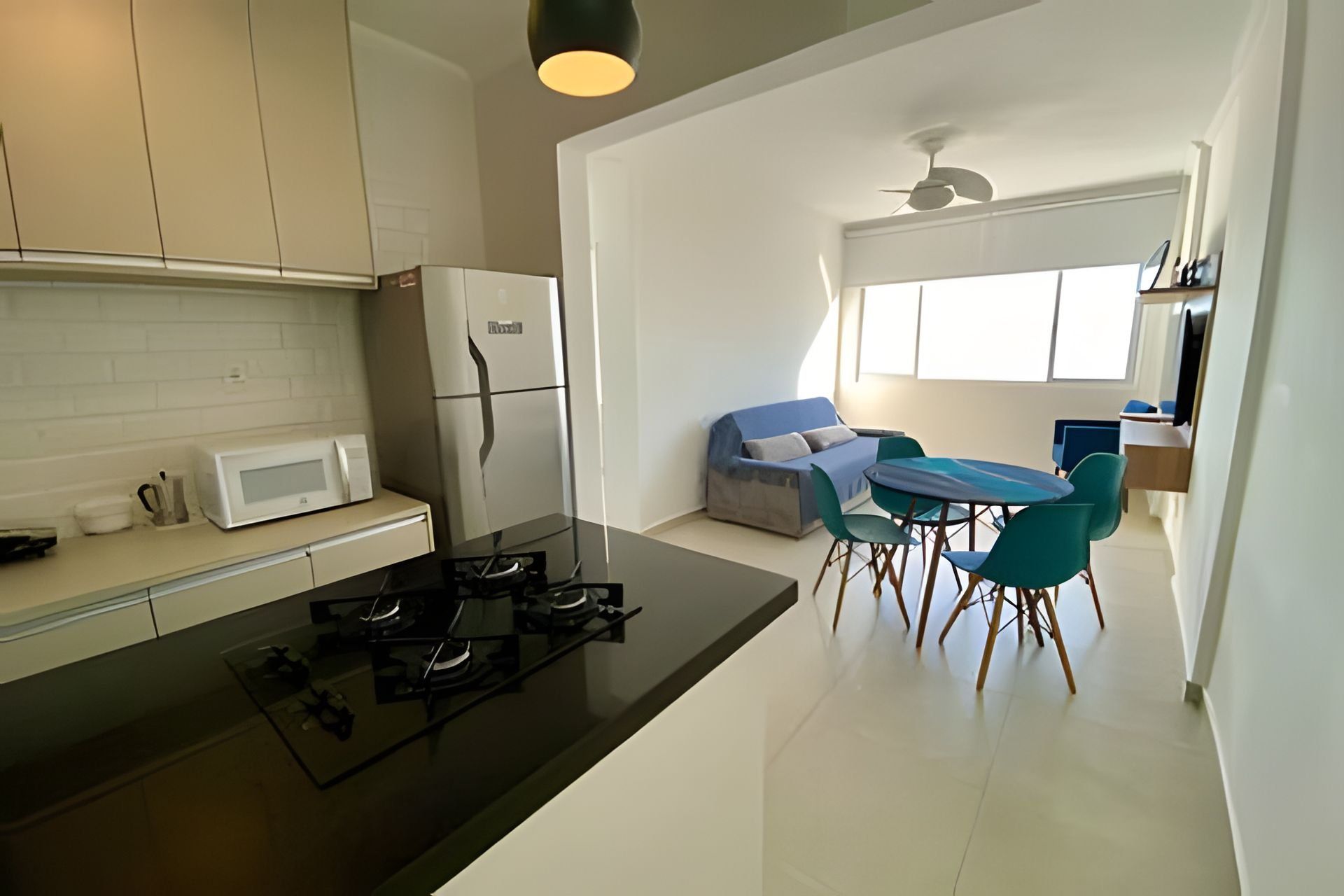 Una cocina moderna con encimera negra, armarios blancos, un refrigerador blanco y una estufa a gas. Hay una zona de comedor con una mesa azul y sillas color turquesa.