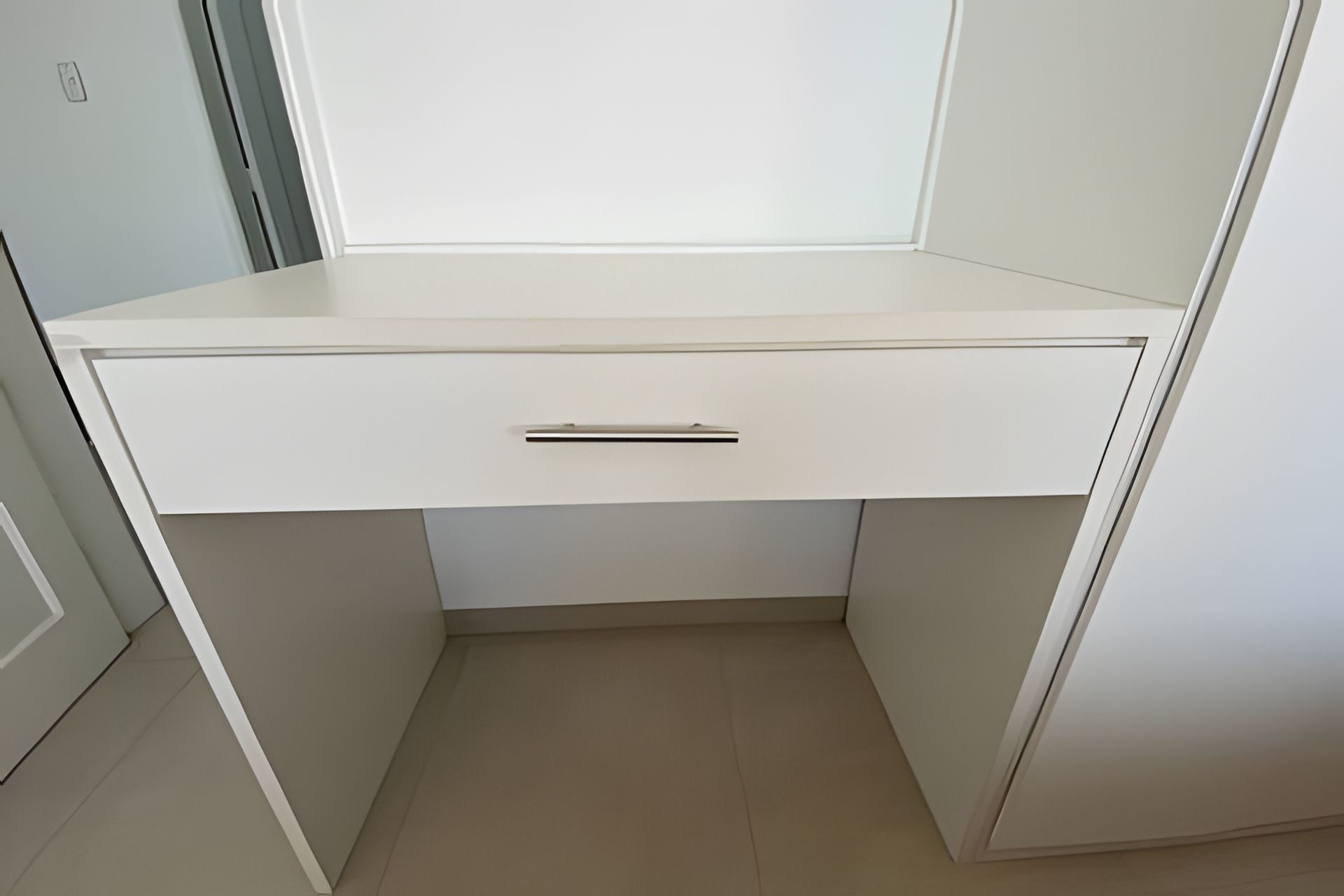 A white desk with a drawer, positioned in a corner of a room with light-colored walls and floor.