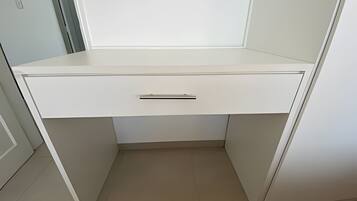 A white desk with a drawer, positioned in a corner of a room with light-colored walls and floor.