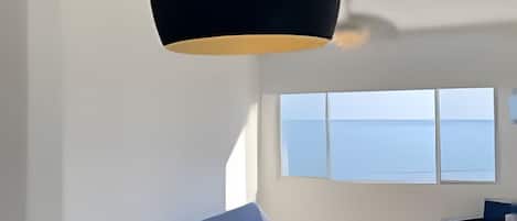 A modern living room with a sofa, a dining table, and a large window offering a view of the sea.