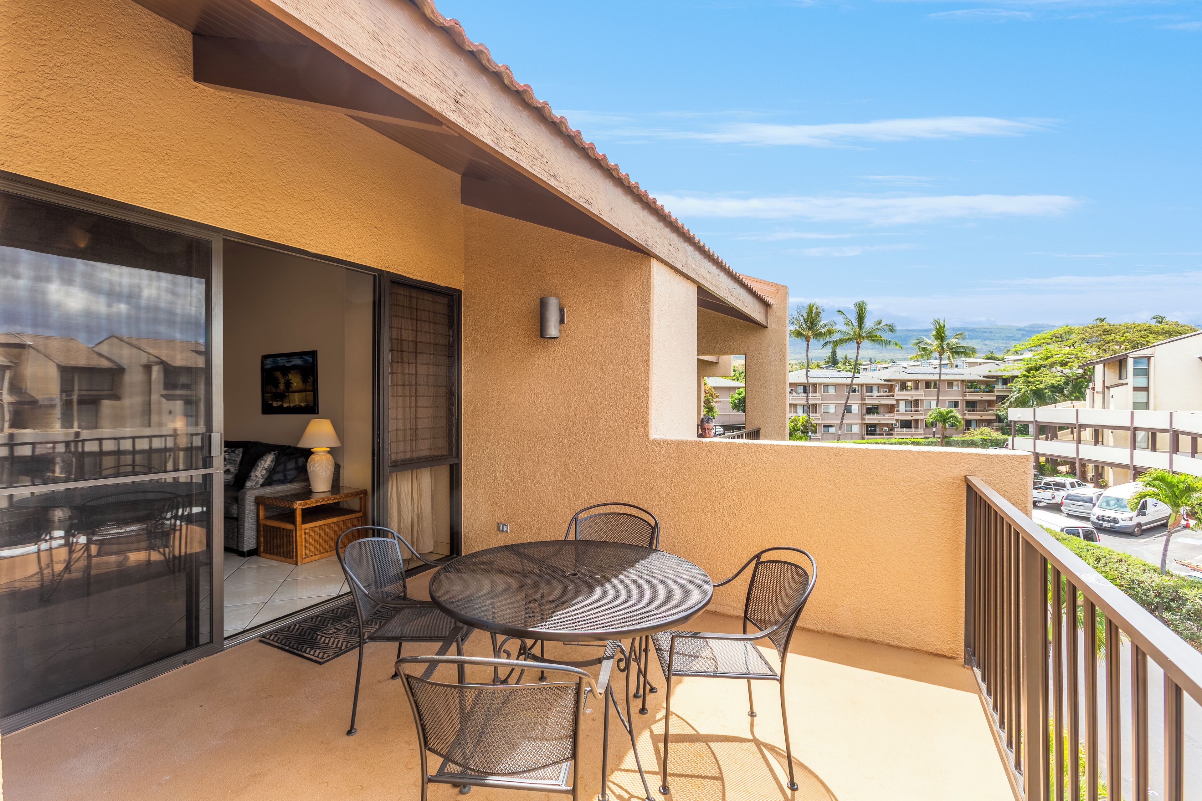 A balcony with a table and chairs, a sliding glass door, and a view of a residential area with palm trees and buildings.