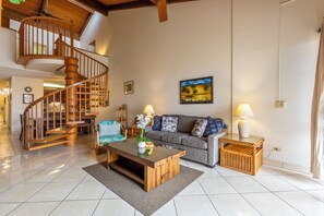 A spacious living area with a wooden staircase, a sofa set, a coffee table, and a painting on the wall. - Kamaole Sands Top Floor 2BR Loft, Ocean View, Mountain View, Beach & Resort Pool (Kihei)