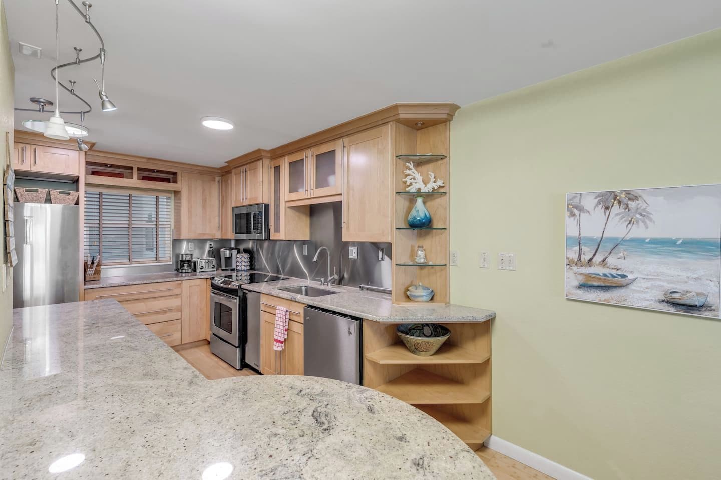A kitchen with granite countertops, stainless steel appliances, wooden cabinets, and a painting of a beach scene.