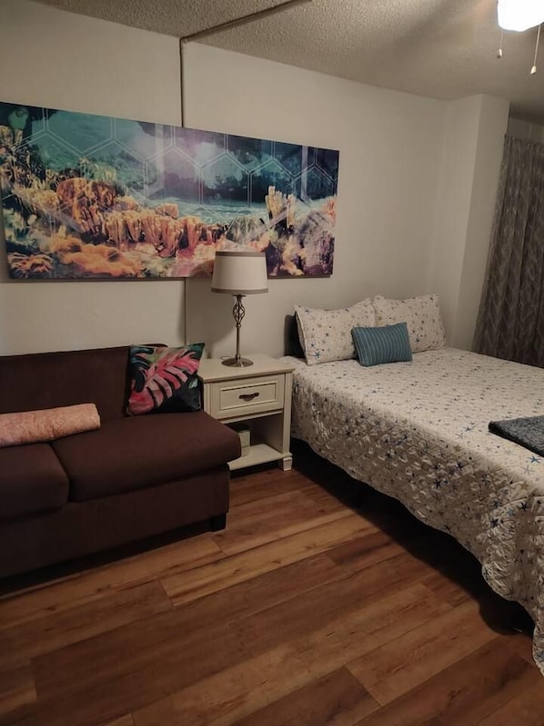 A bedroom with a bed, a nightstand, a lamp, a sofa, and a wall-mounted painting.