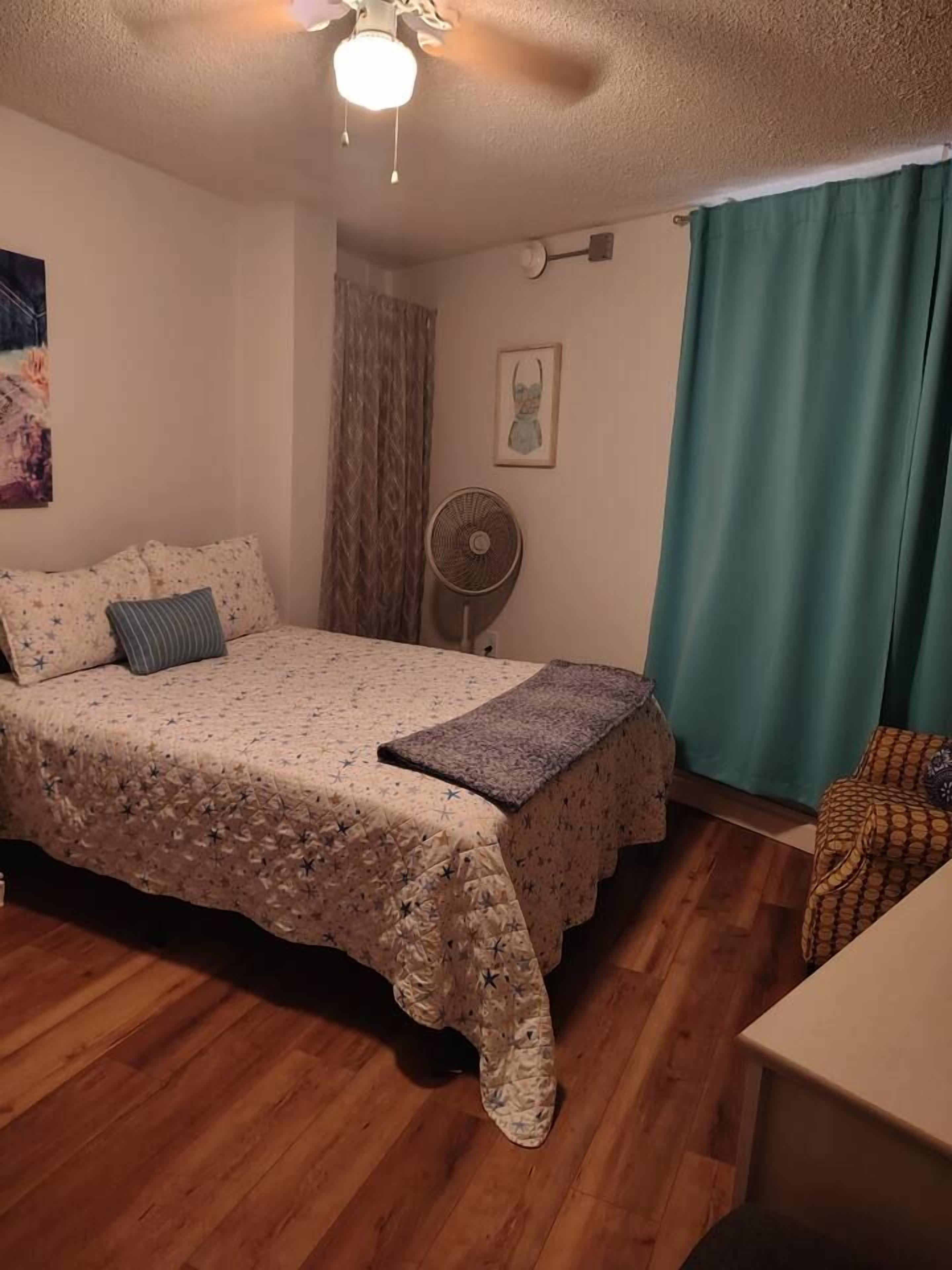 Un dormitorio con una cama, un ventilador, un cuadro en la pared y una ventana con cortinas.