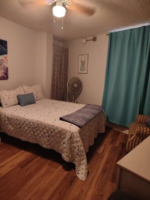 Un dormitorio con una cama, un ventilador, un cuadro en la pared y una ventana con cortinas.