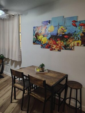 Un comedor con una mesa y sillas de madera, una planta en maceta y una pared con tres cuadros coloridos.