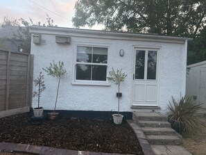 A small white building with two windows and a door, surrounded by a wooden fence and potted plants.