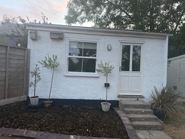 A small white building with two windows and a door, surrounded by a wooden fence and potted plants.