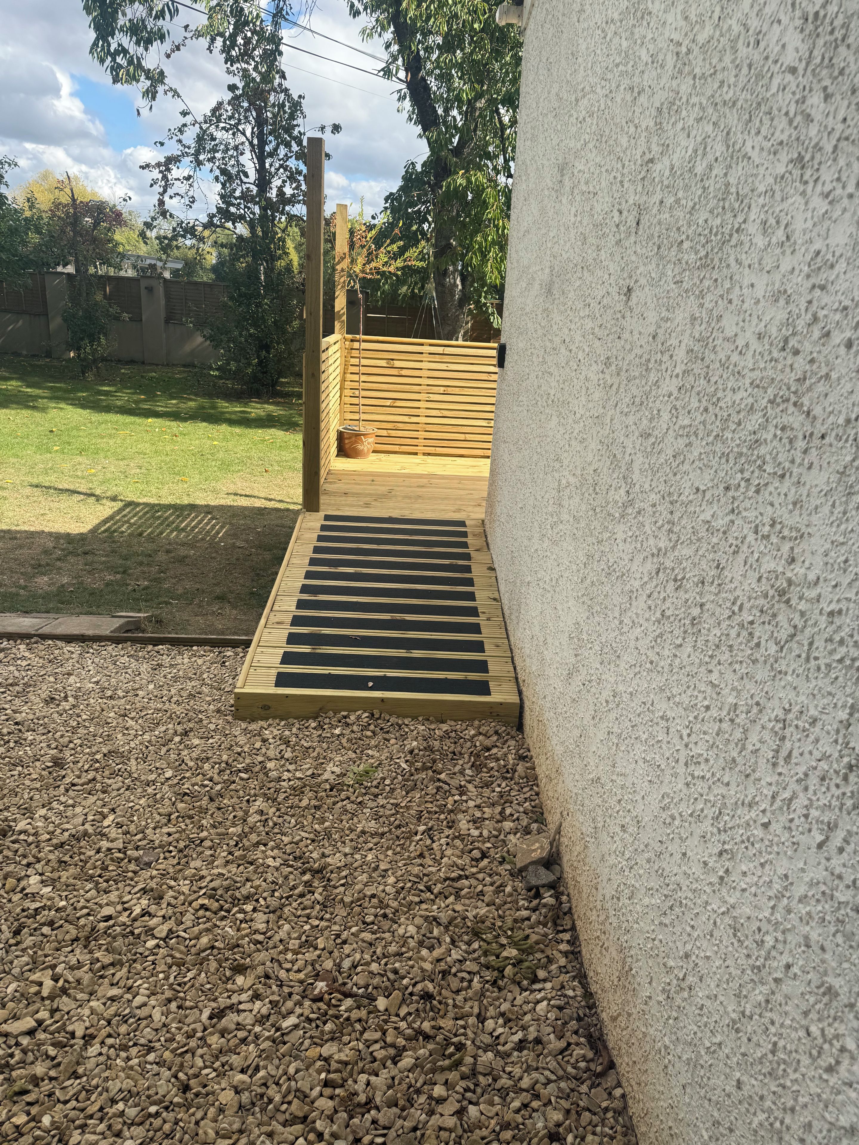 Een houten terras met een metalen rooster dat uitkomt op een grindpad, omgeven door een betonnen muur en een houten omheining.